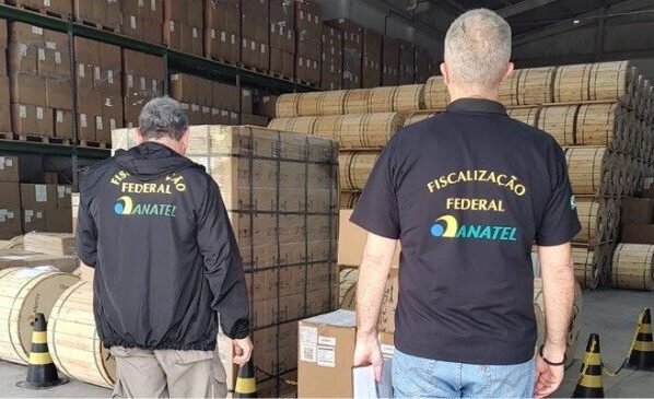 Foto de dois agentes, vistos de costas, em um depósito ou galpão. Eles usam uniformes pretos com o logo da Agência Nacional de Telecomunicações (Anatel) e o texto "Fiscalização Federal ANATEL" em amarelo e azul