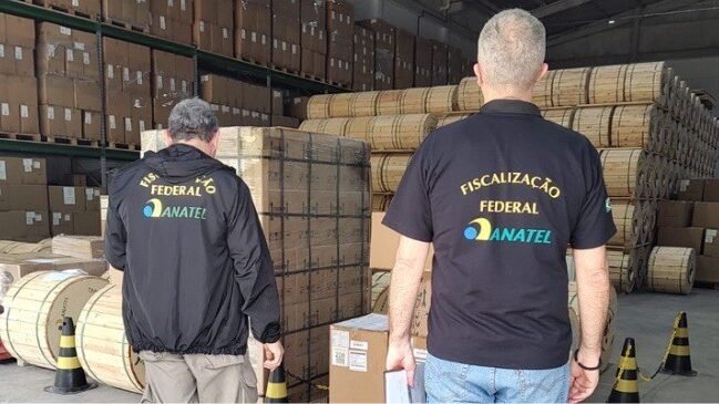 Foto de dois agentes, vistos de costas, em um depósito ou galpão. Eles usam uniformes pretos com o logo da Agência Nacional de Telecomunicações (Anatel) e o texto "Fiscalização Federal ANATEL" em amarelo e azul