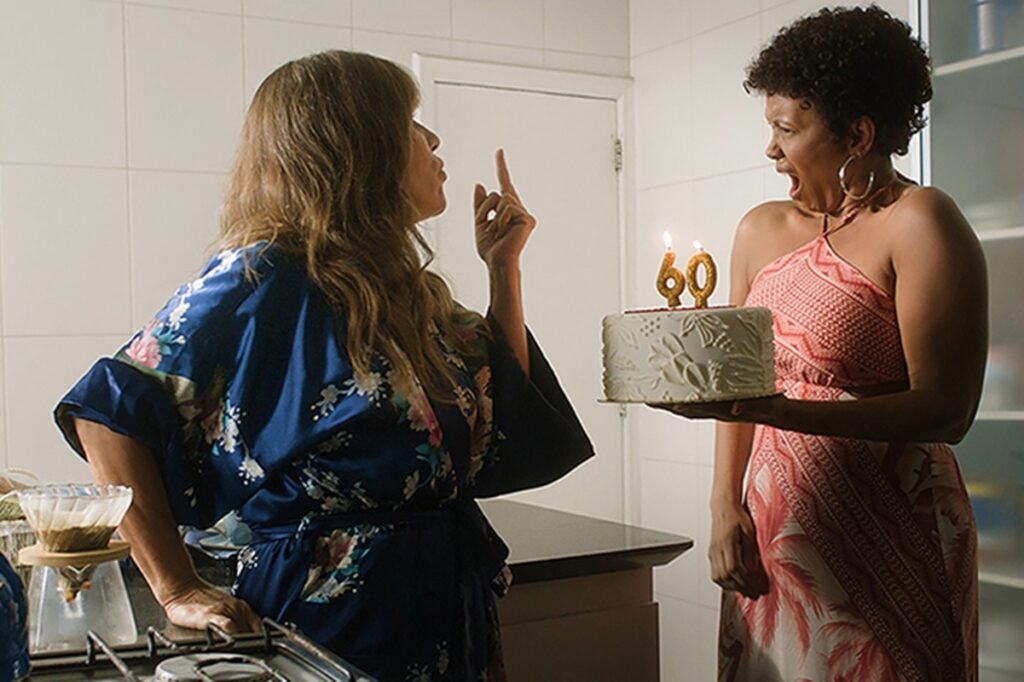 foto colorida de gloria pires, mulher branca de cabelos castanhos comprimos vestindo robe azul estampado, dando dedo para isabel filardis, mulher negra de cabelos curtos cacheados vestida em vestido estampado segurando um bolo, em cena do filme sexa - metrópoles