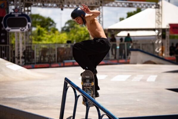 Diversos skatistas executam manobras sobre corrimãos, rampas e bordas de concreto. O céu está azul e o público acompanha das arquibancadas. Os atletas usam roupas leves e capacetes, enquanto deslizam em alta velocidade sob o sol do Parque da Cidade