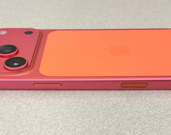 Lateral e parte traseira de um mockup ou conceito do iPhone 17 Pro deitado, mostrando a lateral em cor rosa avermelhada ('rose') com botões, e a parte traseira bipartida em dois tons: o módulo de câmera (três lentes grandes) em rosa avermelhado e o restante do painel traseiro em laranja vibrante, com o logotipo da Apple sutilmente visível.