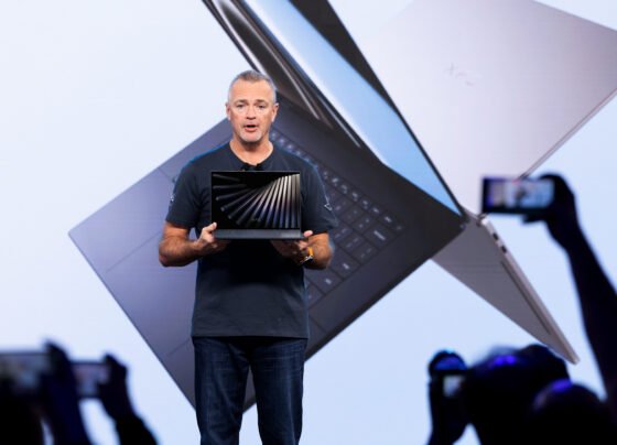 Fotografia colorida mostra o executivo Jeff Clarke no centro de um palco, segurando um notebook aberto. Ele é um homem branco de cabelos curtos grisalhos e veste camiseta azul-marinho e calça jeans. Ao fundo, um telão exibe a imagem ampliada de dois notebooks prata que se cruzam, com a inscrição "XPS" visível em um deles. Em primeiro plano, silhuetas de pessoas na plateia levantam celulares para fotografar o momento. O ambiente tem iluminação clara e tons azulados.