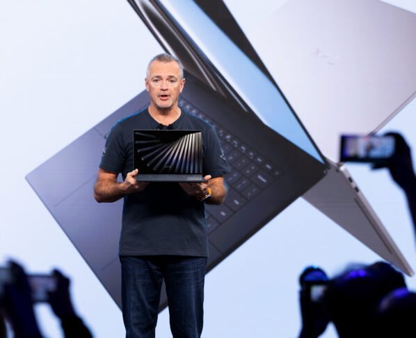 Fotografia colorida mostra o executivo Jeff Clarke no centro de um palco, segurando um notebook aberto. Ele é um homem branco de cabelos curtos grisalhos e veste camiseta azul-marinho e calça jeans. Ao fundo, um telão exibe a imagem ampliada de dois notebooks prata que se cruzam, com a inscrição "XPS" visível em um deles. Em primeiro plano, silhuetas de pessoas na plateia levantam celulares para fotografar o momento. O ambiente tem iluminação clara e tons azulados.