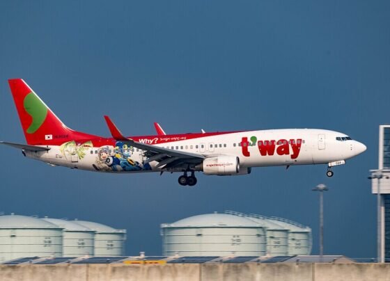 Fotografia em plano médio de um avião da companhia sul-coreana T'way Air em fase de pouso. A aeronave é branca com a cauda vermelha e traz a inscrição "t'way" em letras grandes e vermelhas na fuselagem. Na parte traseira, há uma ilustração colorida de personagens de desenho animado e a pergunta "Why?". O céu ao fundo é azul-escuro e, na parte inferior, veem-se tanques de combustível brancos e o topo de um edifício de aeroporto.