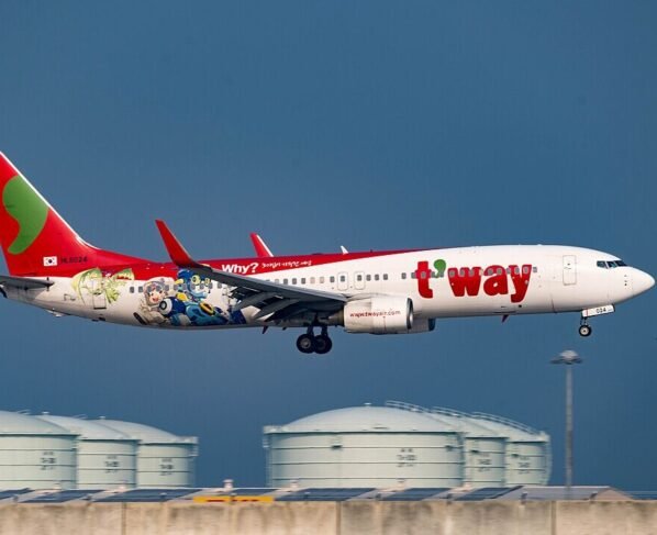 Fotografia em plano médio de um avião da companhia sul-coreana T'way Air em fase de pouso. A aeronave é branca com a cauda vermelha e traz a inscrição "t'way" em letras grandes e vermelhas na fuselagem. Na parte traseira, há uma ilustração colorida de personagens de desenho animado e a pergunta "Why?". O céu ao fundo é azul-escuro e, na parte inferior, veem-se tanques de combustível brancos e o topo de um edifício de aeroporto.