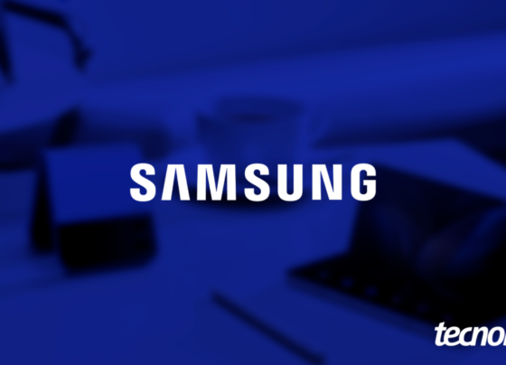 Imagem mostra a palavra "SAMSUNG" sendo exibida no centro, em letras brancas e maiúsculas. O fundo, em tom azul escuro, mostra elementos desfocados que sugerem um ambiente de escritório. Na parte inferior direita, o logotipo do "Tecnoblog" é visível.