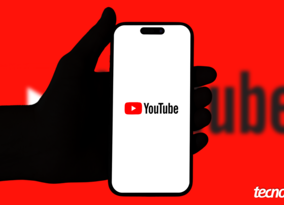 Mão segurando um celular que exibe o YouTube, com um fundo de cor vermelha. Na parte inferior direita, está o logotipo do "tecnoblog".