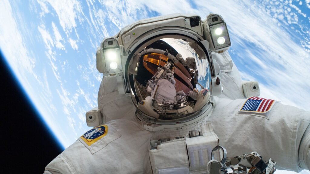 Selfie em close de um astronauta em traje espacial branco durante caminhada espacial. O visor do capacete reflete a estrutura da estação e parte do planeta Terra. Duas luzes brancas intensas estão acesas acima do visor. No braço esquerdo, há uma bandeira dos Estados Unidos e, no direito, um emblema circular colorido. O fundo exibe a curvatura da Terra com tons de azul intenso e nuvens brancas sobre o espaço negro. Na parte inferior, veem-se ferramentas e partes metálicas do traje.