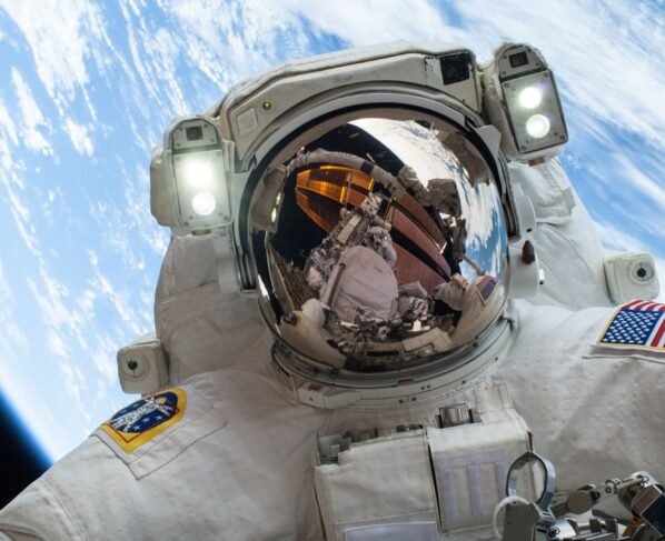 Selfie em close de um astronauta em traje espacial branco durante caminhada espacial. O visor do capacete reflete a estrutura da estação e parte do planeta Terra. Duas luzes brancas intensas estão acesas acima do visor. No braço esquerdo, há uma bandeira dos Estados Unidos e, no direito, um emblema circular colorido. O fundo exibe a curvatura da Terra com tons de azul intenso e nuvens brancas sobre o espaço negro. Na parte inferior, veem-se ferramentas e partes metálicas do traje.