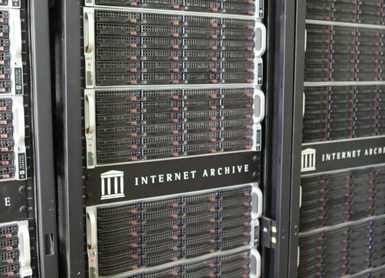 Fotografia em plano médio e ângulo diagonal mostra três racks de servidores pretos alinhados lateralmente. Os equipamentos possuem diversas gavetas horizontais com frentes gradeadas e pequenas luzes indicadoras acesas em tons de azul e vermelho. No centro de cada rack, há uma placa preta com o logotipo de um templo grego e o texto "INTERNET ARCHIVE" em letras brancas e maiúsculas. Nas laterais, veem-se etiquetas brancas numeradas e alças de metal prateadas para manuseio.